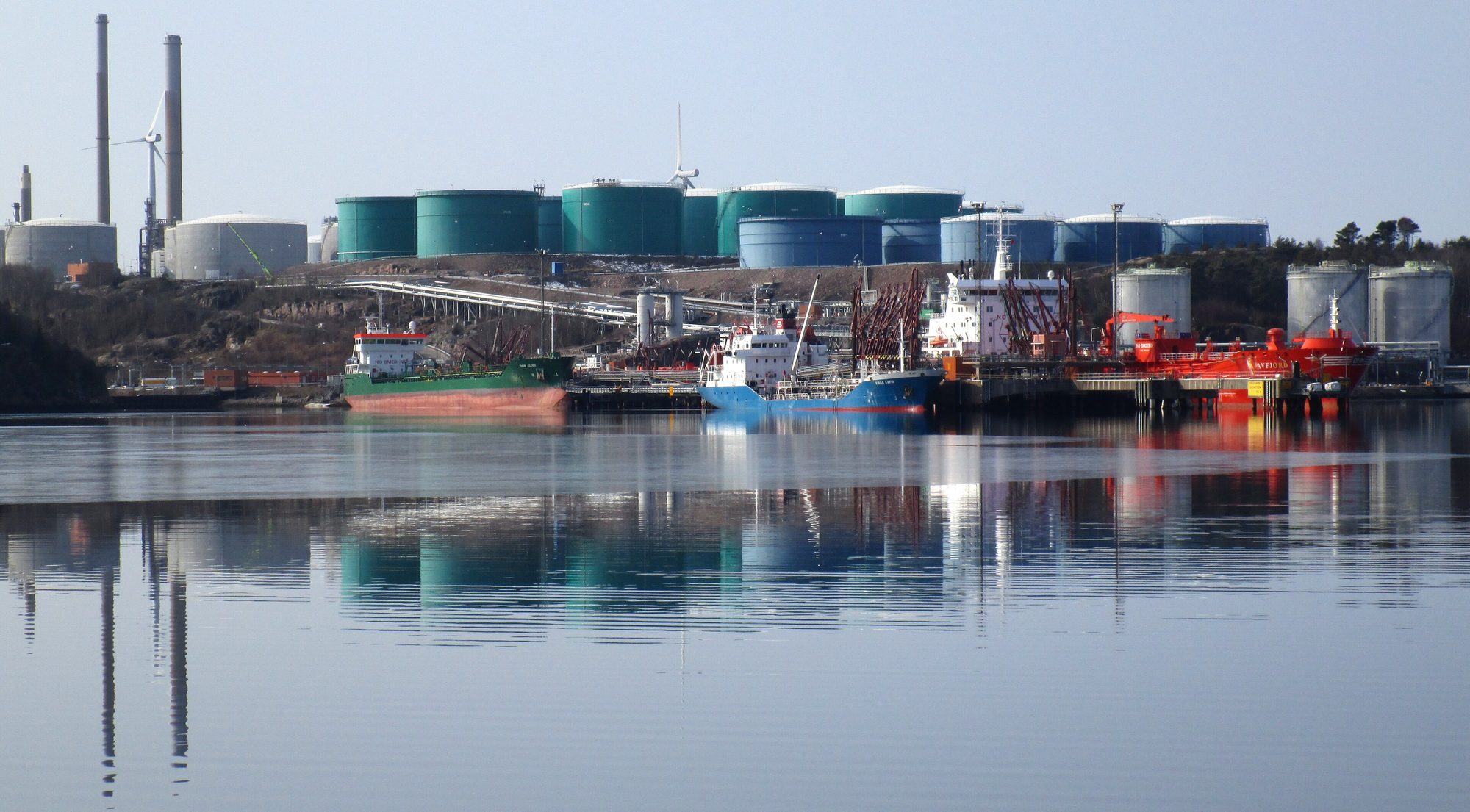 Tankers afgemeerd aan een steiger van een olieterminal met opslagtanks erachter, weerspiegeld in kalm water